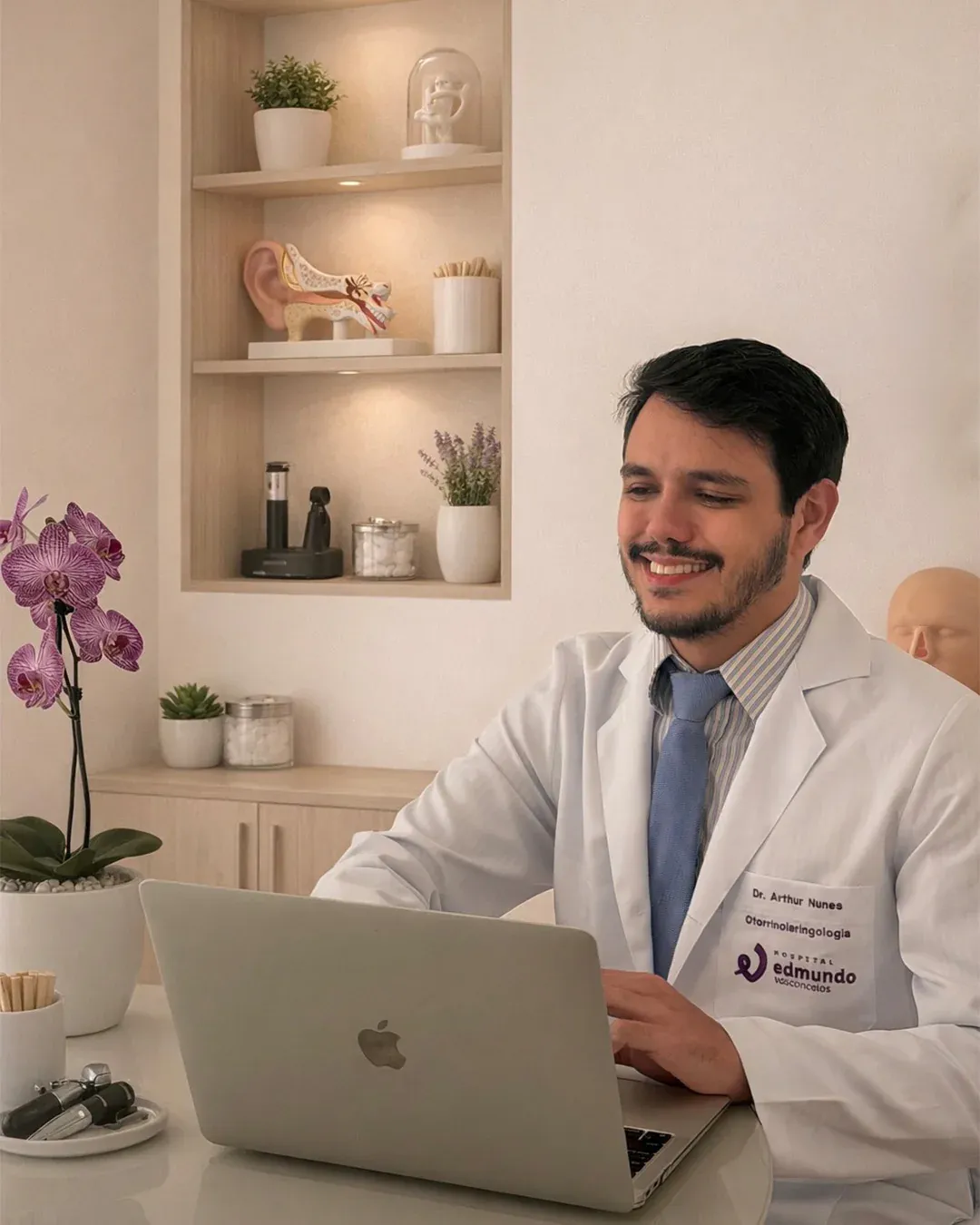 Dr. Arthur Nunes em seu consultório, sorrindo enquanto trabalha no computador, com modelo anatômico de ouvido ao fundo.